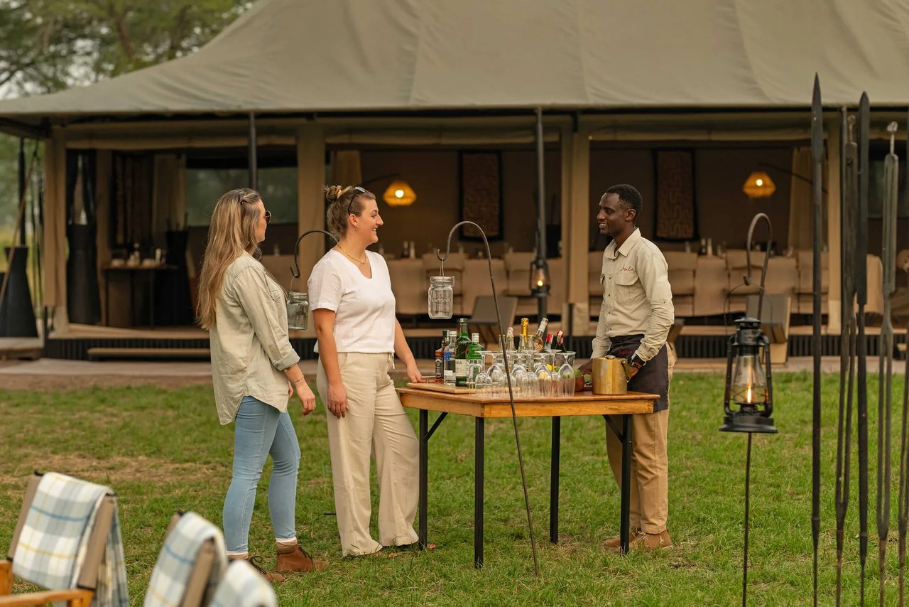 A camp host in a dark uniform serves welcome drinks to guests seated at Ubuntu Camp