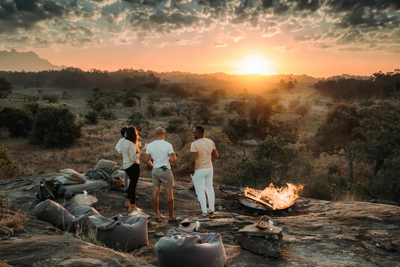 Travellers gather at a firepit on the rocks as the sun drops behind the hills