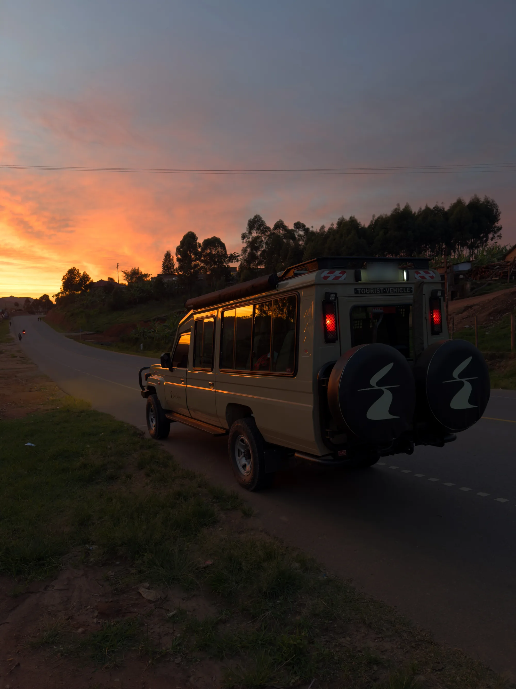 A Sail Safaris DMC vehicle parked at the roadside at dawn on the road to Queen Elizabeth National Park in Uganda, the Equator Fold symbol on both spare-wheel covers and the road curving away towards the sunrise