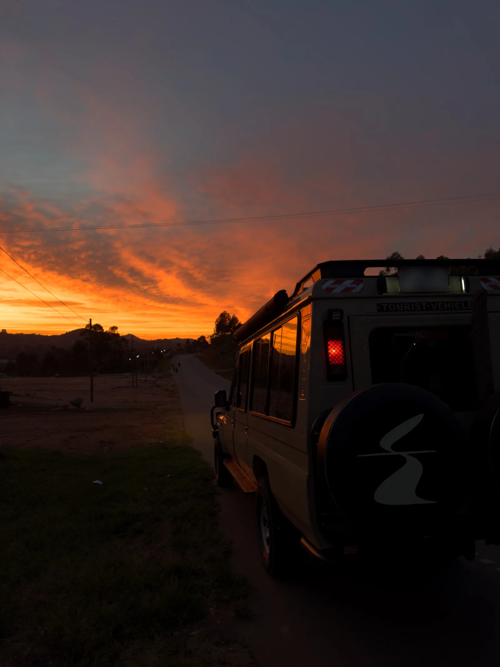 The rear of a Sail Safaris DMC vehicle at the roadside at dawn on the road to Queen Elizabeth National Park in Uganda, the Equator Fold symbol on its spare-wheel cover and the road running towards the hills under an orange sky