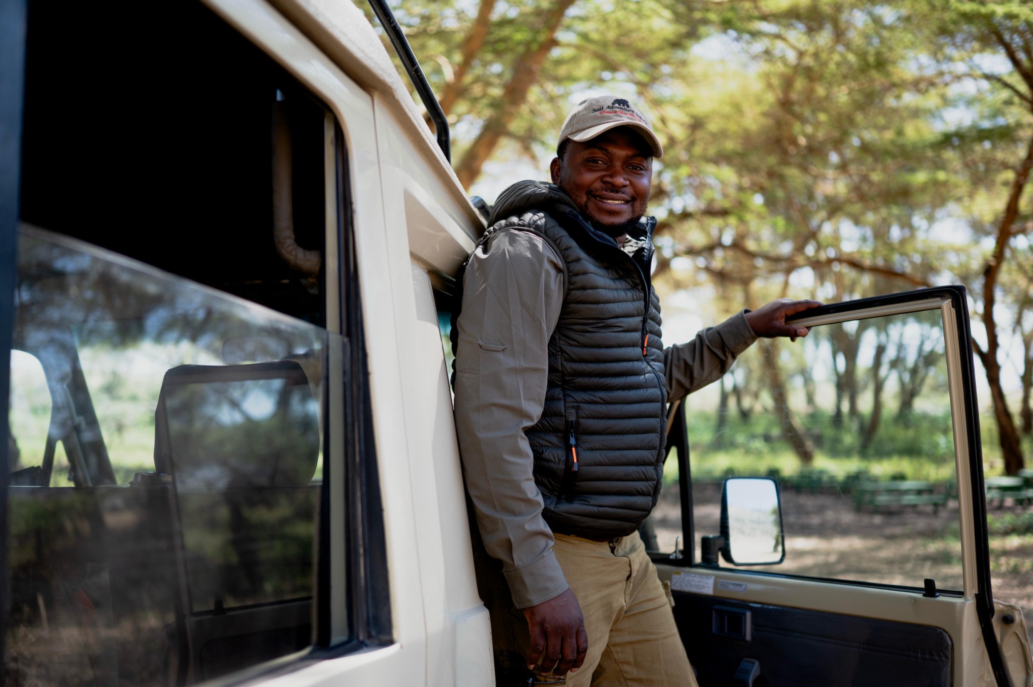 A Sail guide in a gilet standing at the open door of a safari vehicle, savannah behind