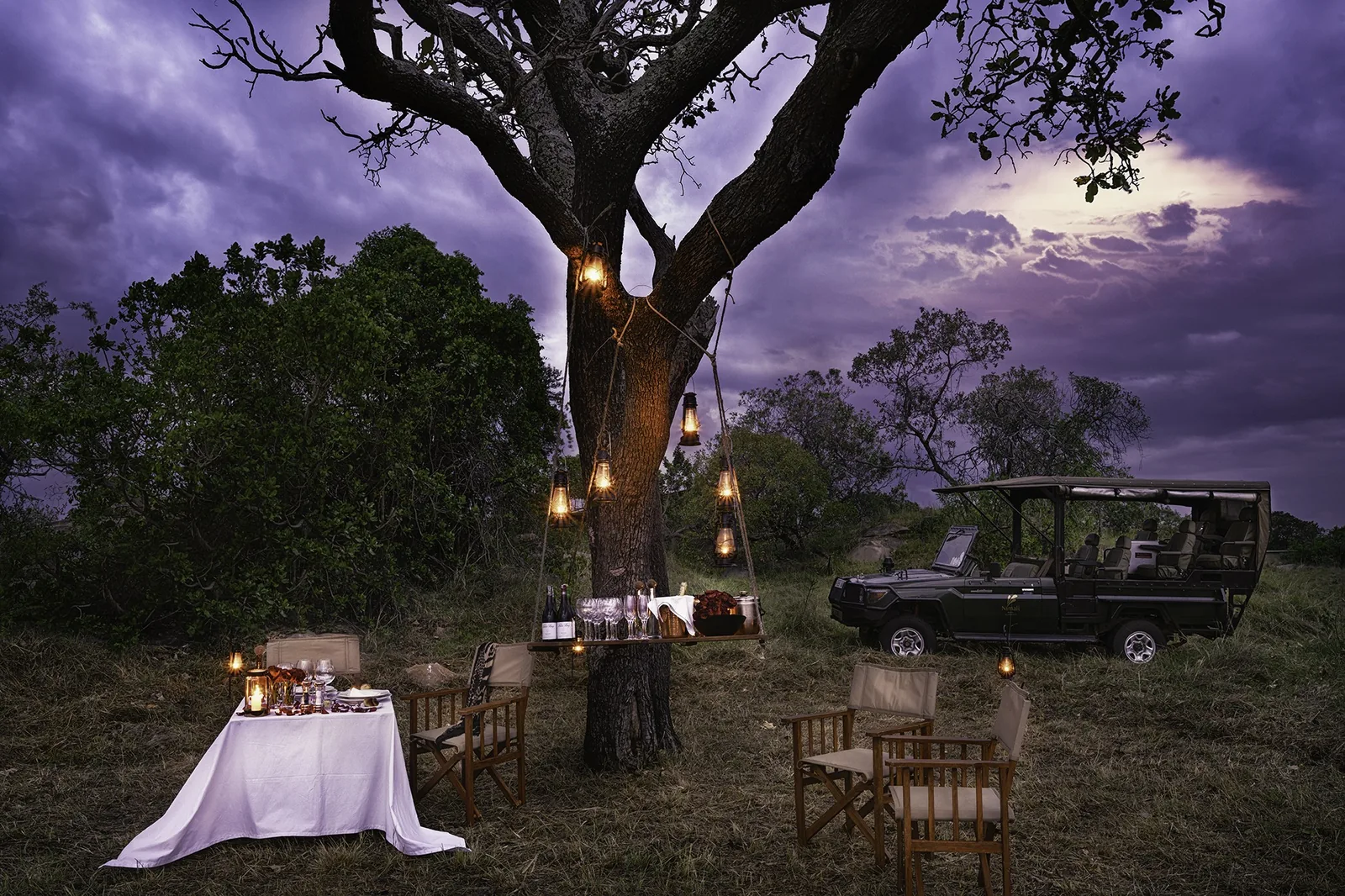 A lantern-lit dinner waits under a tree beside the game-drive vehicle at dusk