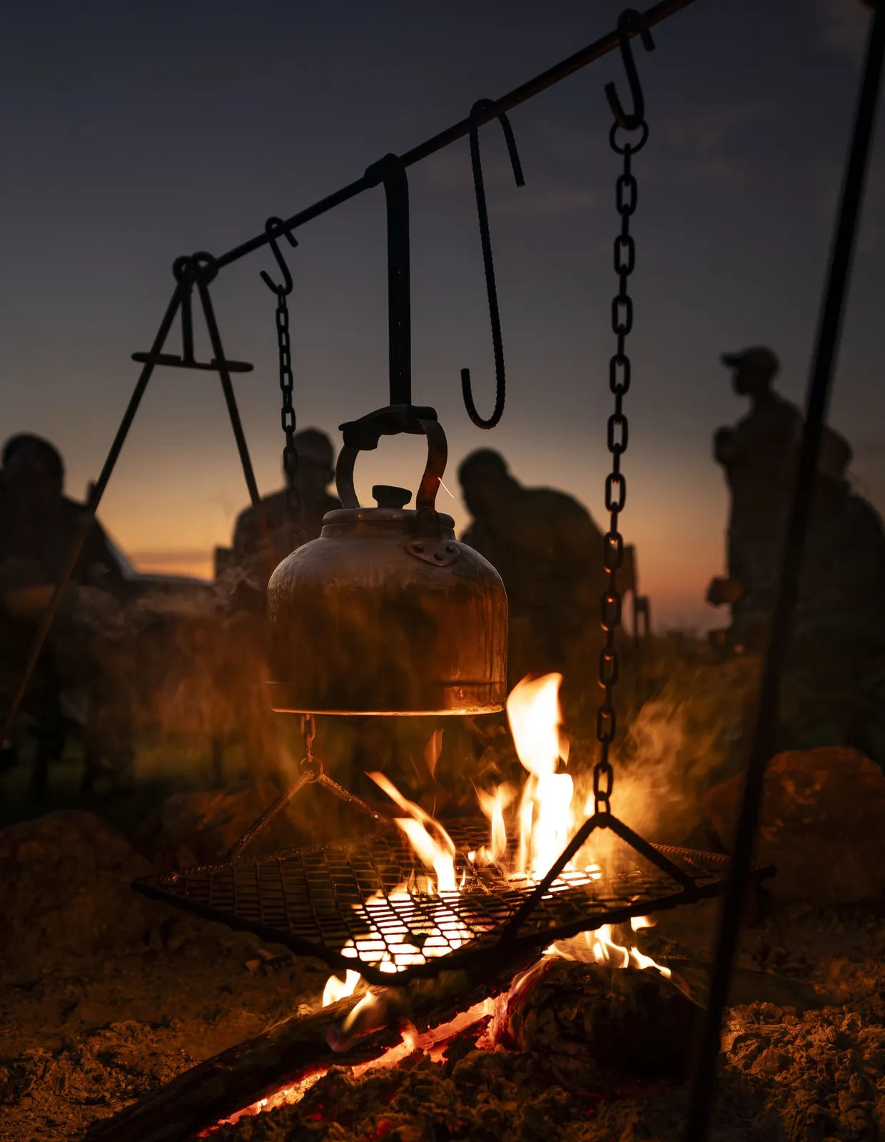 A kettle hangs over a camp fire as dusk silhouettes gather behind