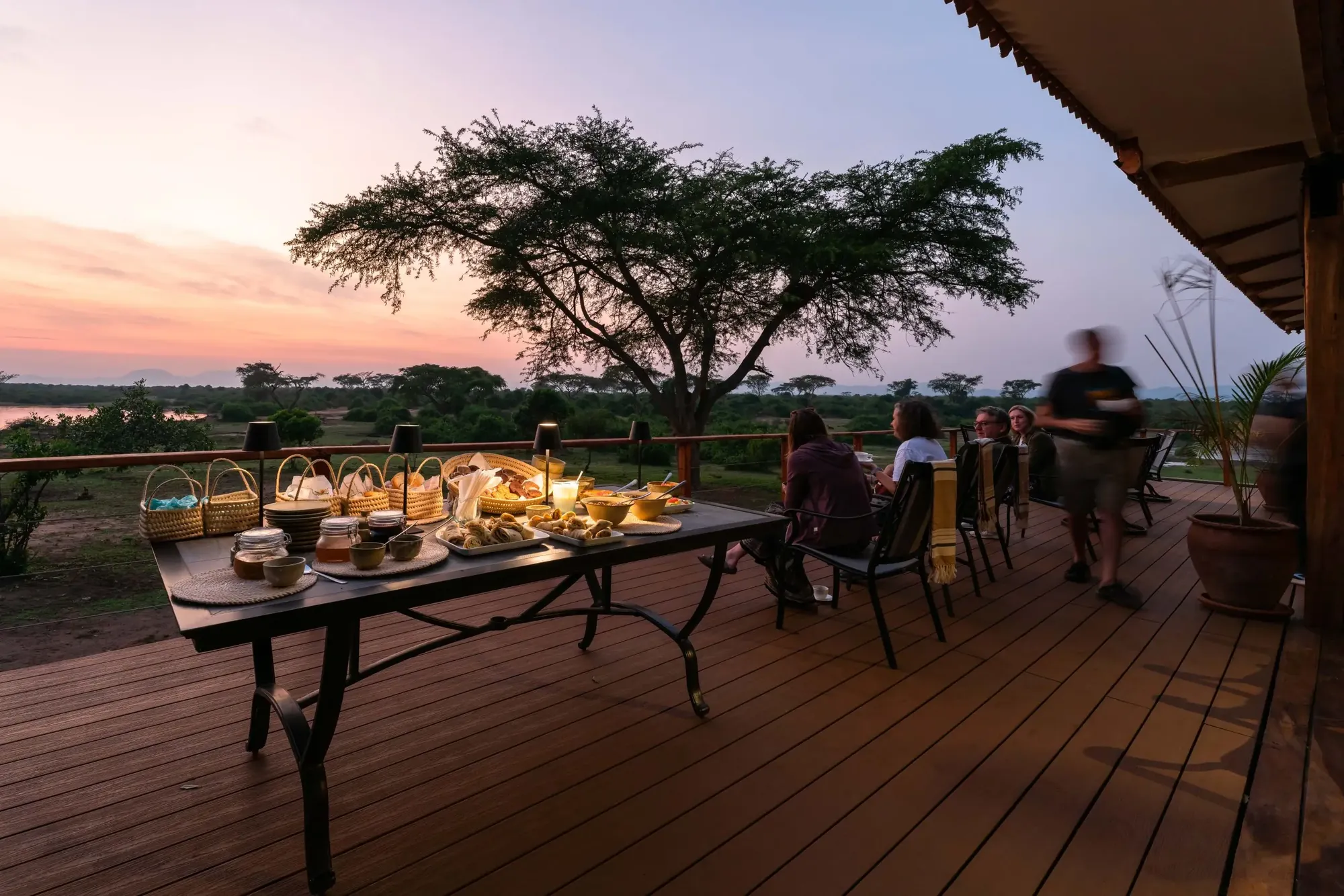 A linen breakfast table sits at the river's edge at sunrise in Queen Elizabeth National Park, mist on the water.