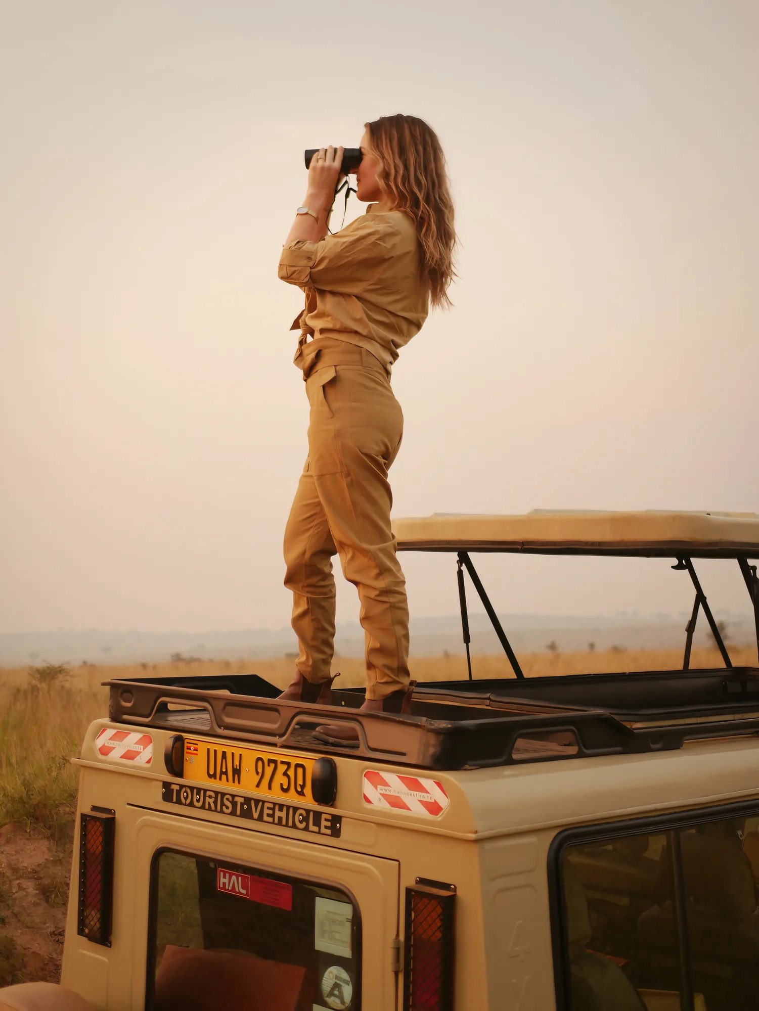A guest stands through the roof hatch of a safari vehicle in Uganda, scanning the country beyond the track.