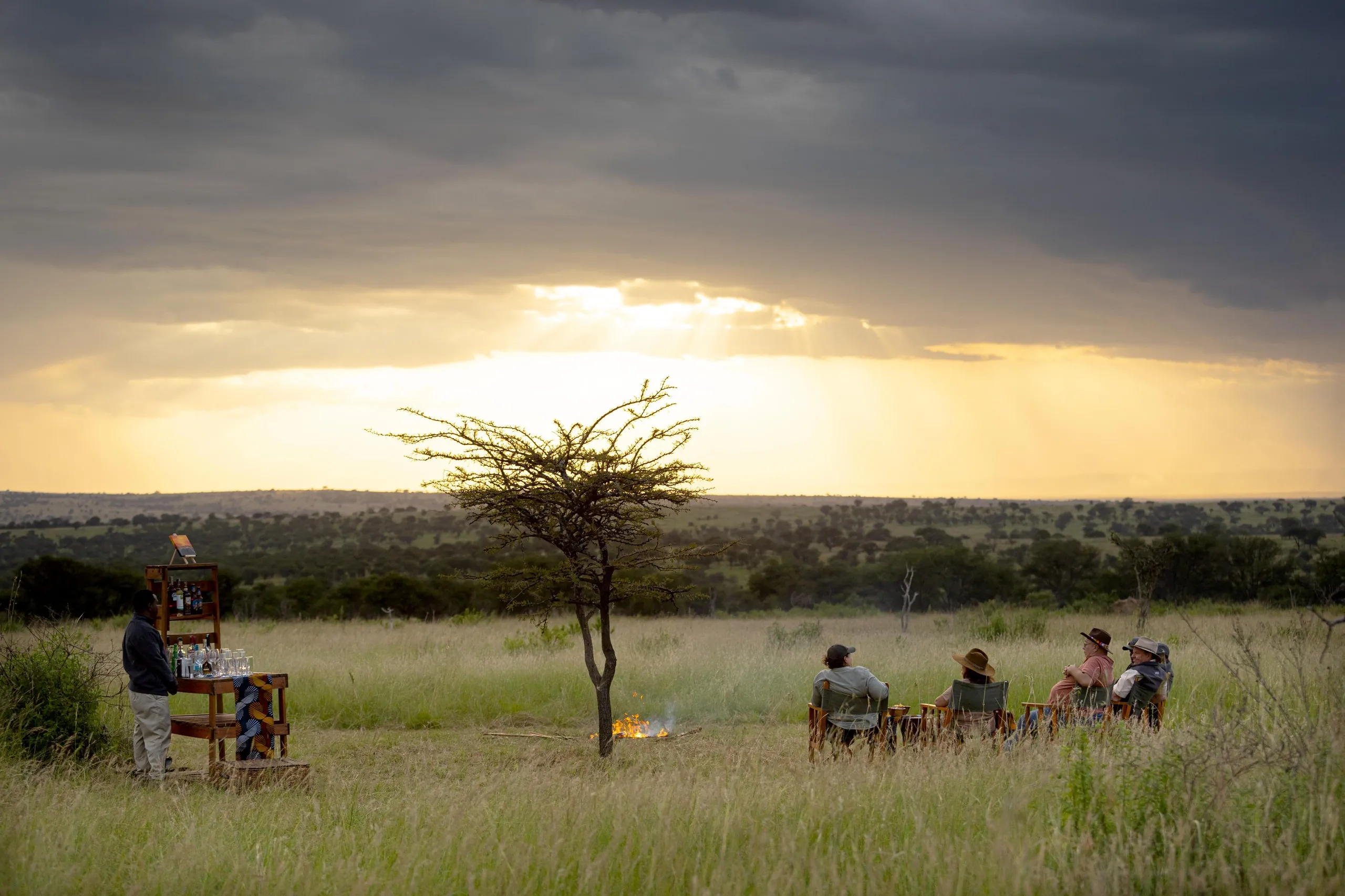 A storm-gold sky breaks over a Mara sundowner fire beneath a lone acacia