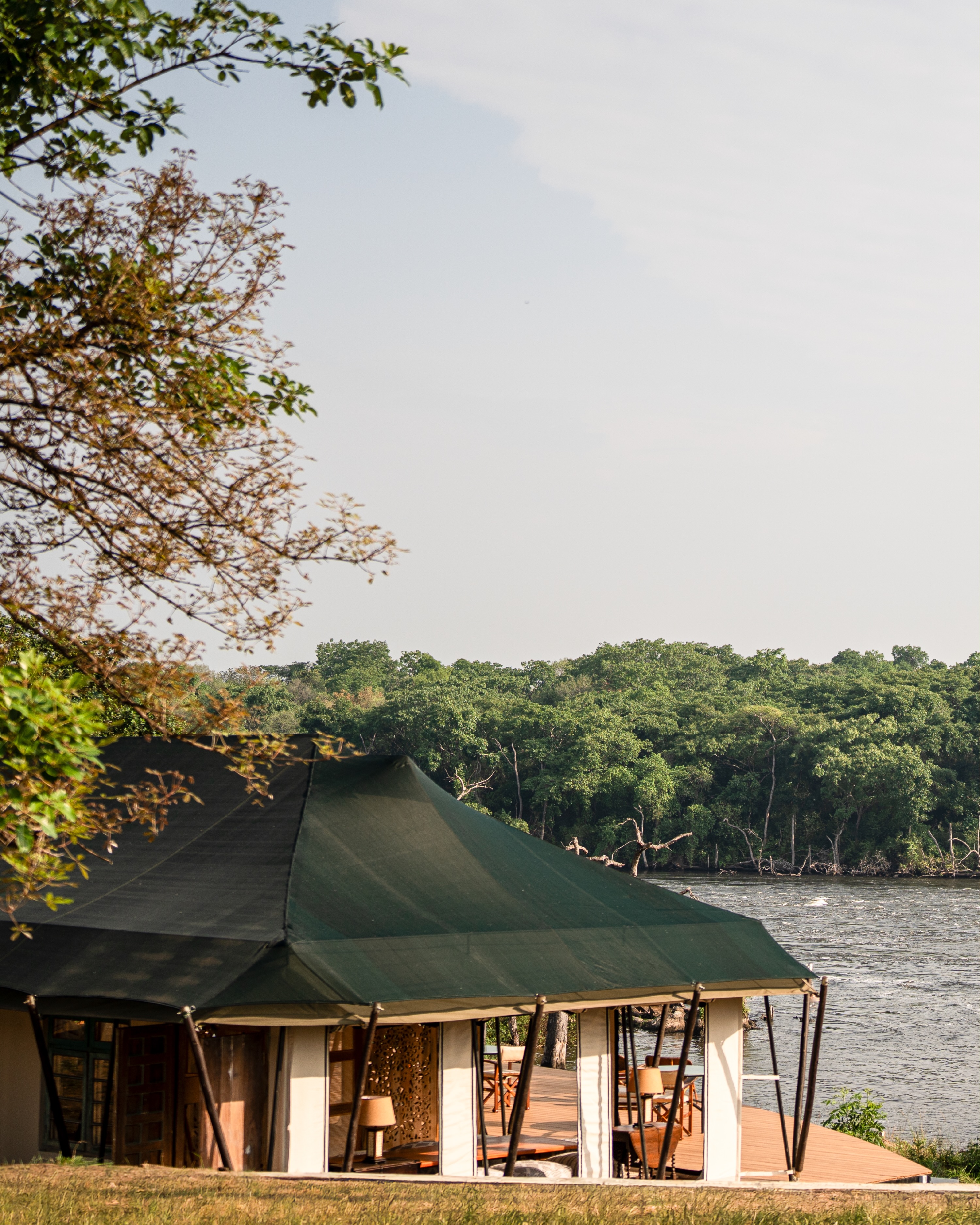 An open-sided tent under a green canvas roof at Kulu Ora, on a lawn beside the Nile, with forest on the far bank