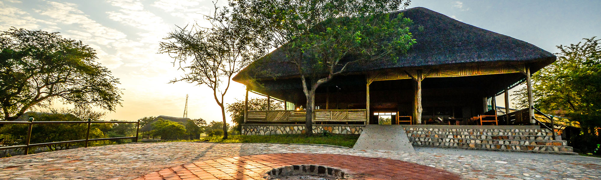 The thatched main building of Twiga Safari Lodge on a stone base, beyond a paved courtyard with a fire pit, in low sun