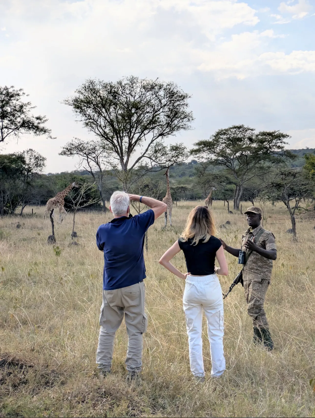 An armed ranger in green walks ahead of guests on a grassy plain, rifle slung, scanning the ground ahead.