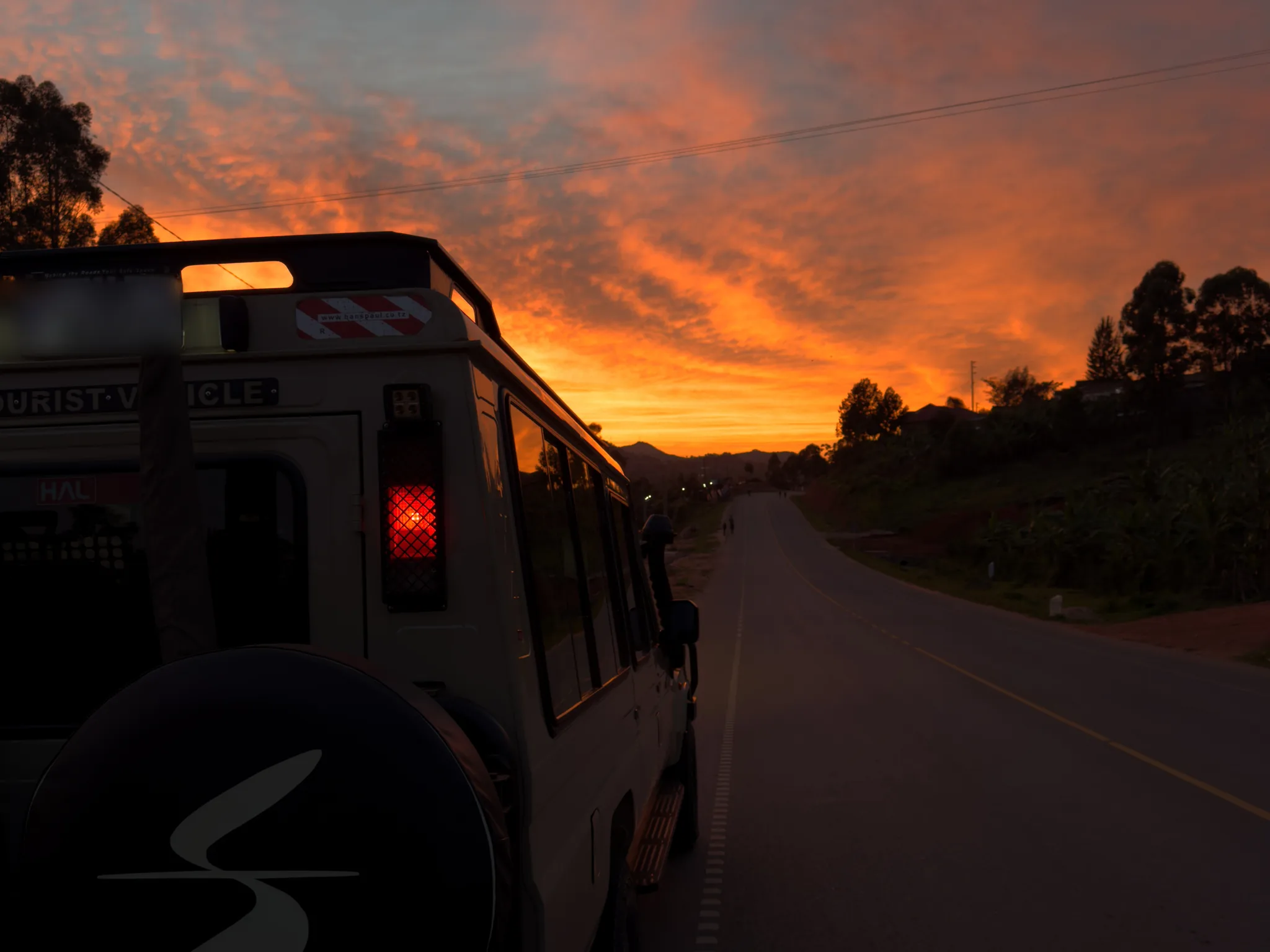 The side of a Sail Safaris DMC vehicle at dawn on the road to Queen Elizabeth National Park, the tarmac running towards hills under an orange sky