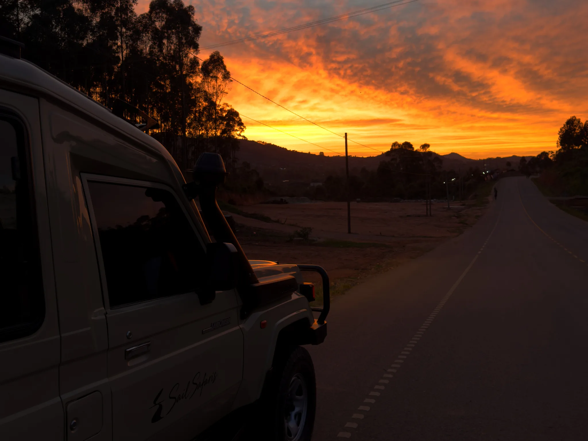 The front of a Sail Safaris DMC vehicle at the roadside at dawn on the road to Queen Elizabeth National Park, which also leads to Bwindi's Buhoma sector, the tarmac running towards hills under an orange sky