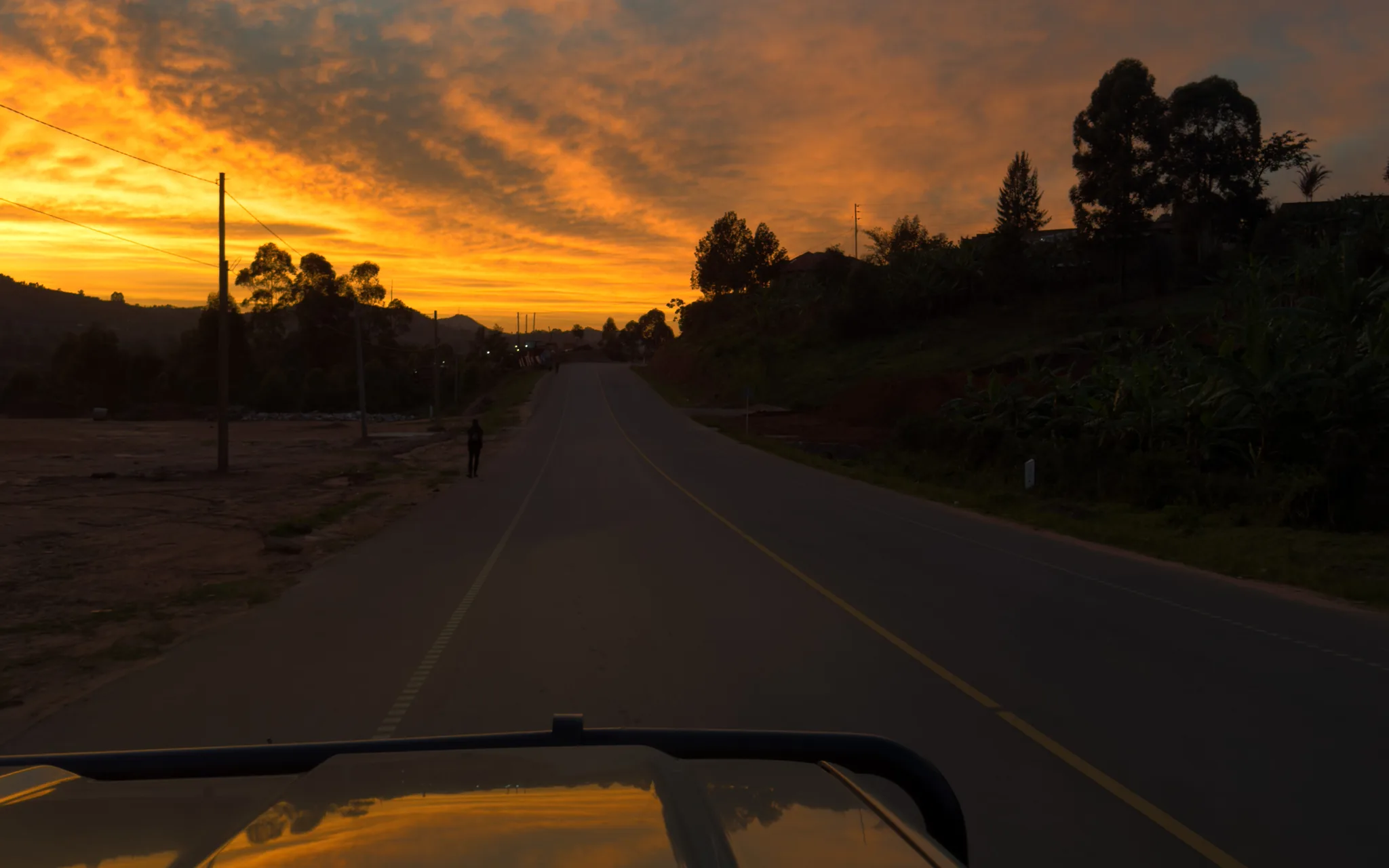 The view over the bonnet of a safari vehicle along the road to Queen Elizabeth National Park at dawn, a walker on the shoulder and the sky orange above the hills