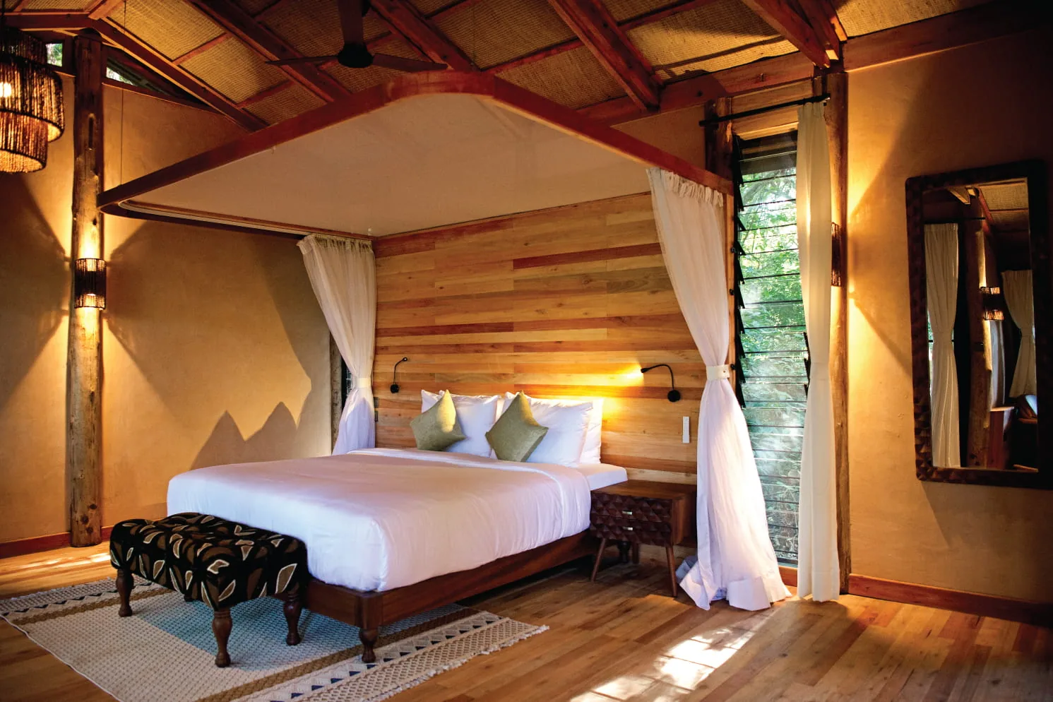 A canopied bed beneath a thatched roof at Entebbe Forest Lodge, with forest light through the louvres